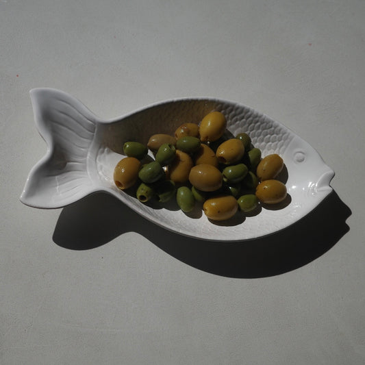 A white ceramic fish-shaped dish containing green and yellow olives, set against a plain light gray background with a strong shadow.