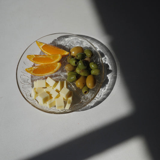 A clear glass plate with embossed floral detailing, holding cubed cheese, green and yellow olives, and orange slices, set on a white surface under direct sunlight with a strong shadow.