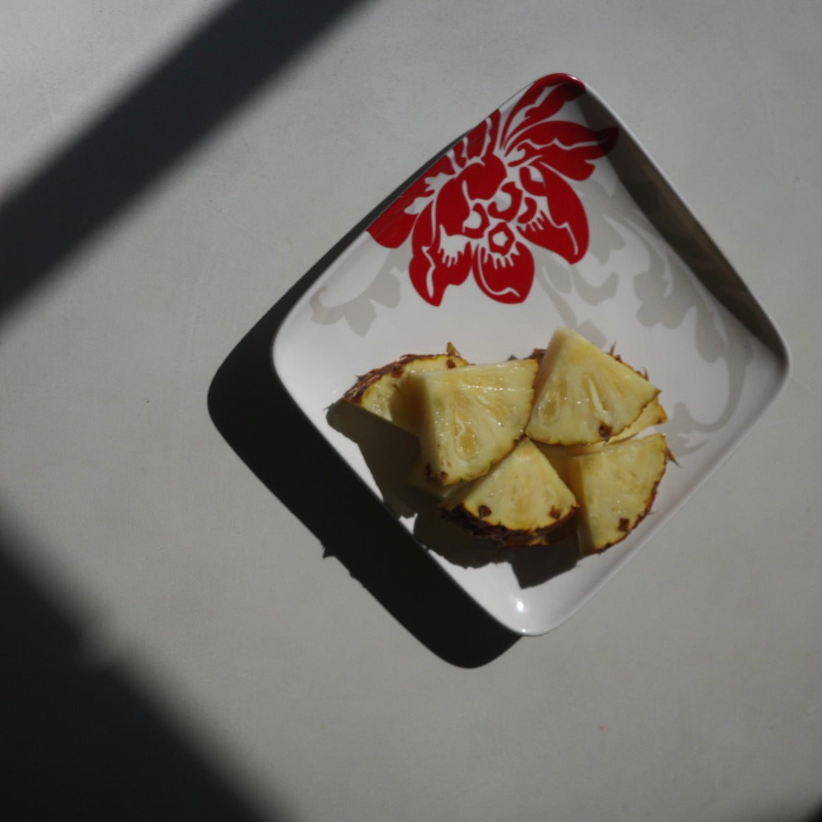 Four slices of pineapple on a square white plate with a red floral pattern, set on a light gray surface under sharp sunlight with a strong shadow.
