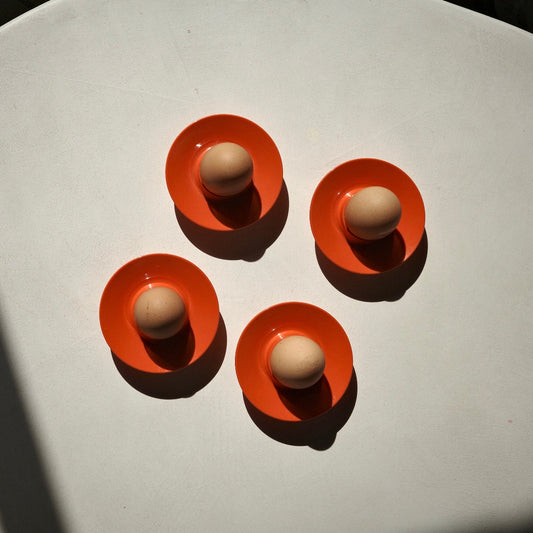 Four orange plastic egg holders arranged in a square on a white surface, each holding a small beige egg under harsh direct sunlight.