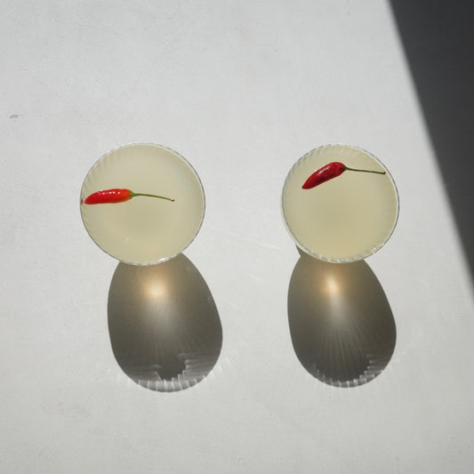 Two clear glass coupe-style cocktails glasses filled with a pale yellow liquid and each containing a small red chili pepper, photographed on a white surface with strong sunlight and shadows.