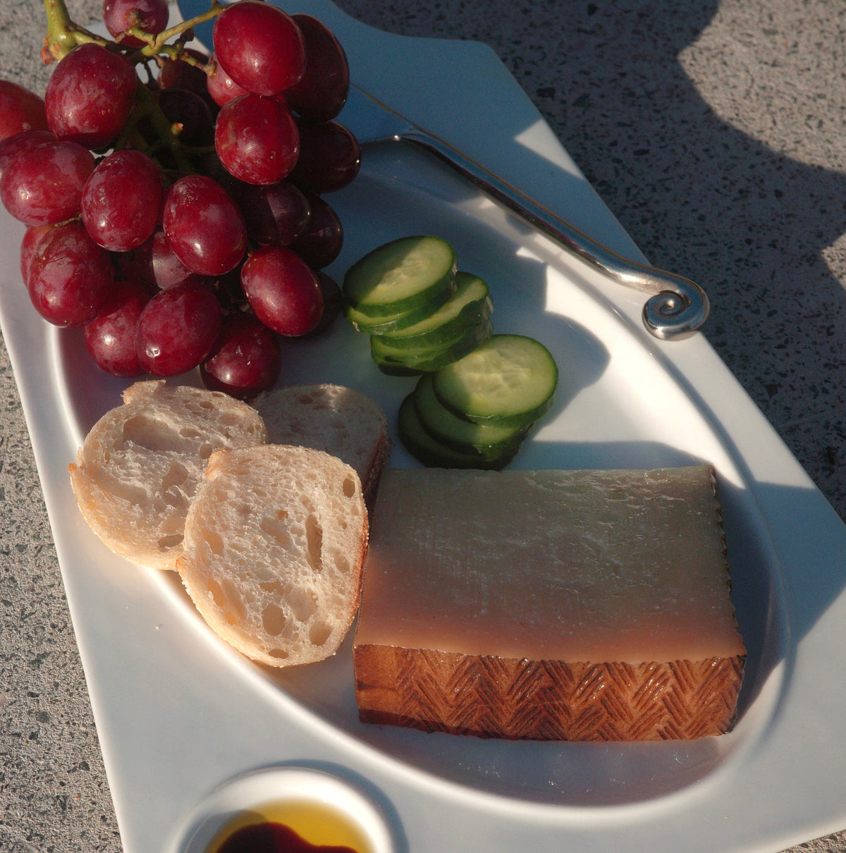 A white serving platter outdoors on a stone surface, arranged with red grapes, sliced bread, cucumber slices, a wedge of cheese. A metal utensil spiral ended knife on platter.