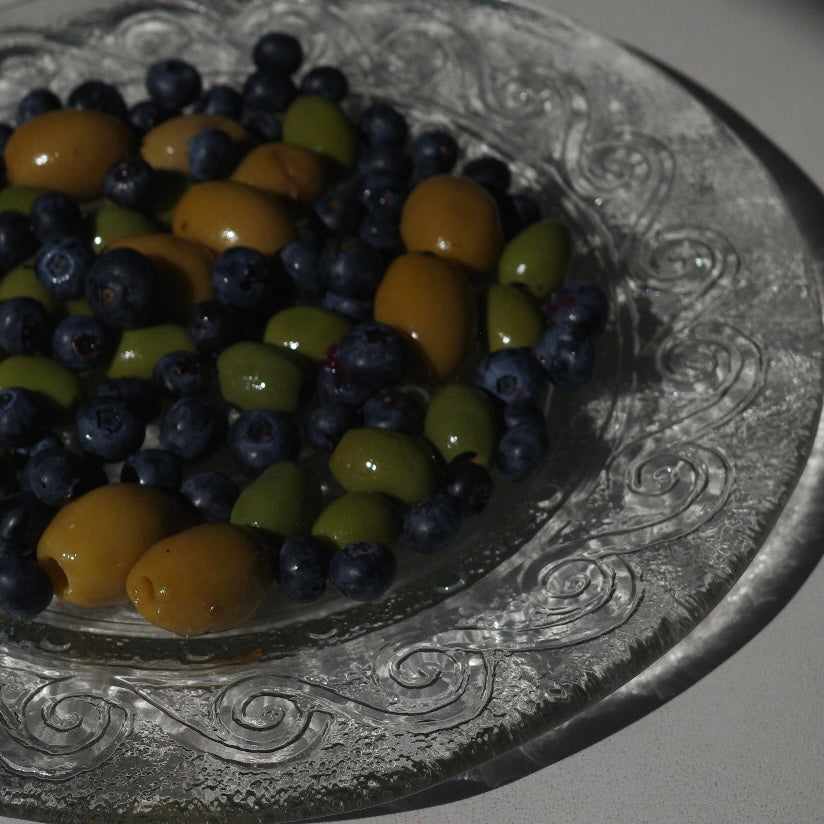 A close-up of mixed berries and green olives arranged on a decorative clear glass plate with an embossed swirl pattern.