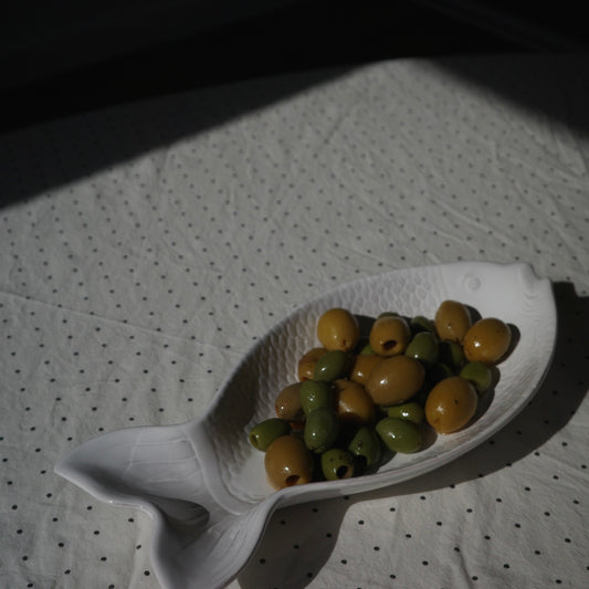 A white fish-shaped ceramic dish holding green and yellow olives, placed on a white polka dot tablecloth under dramatic shadow.