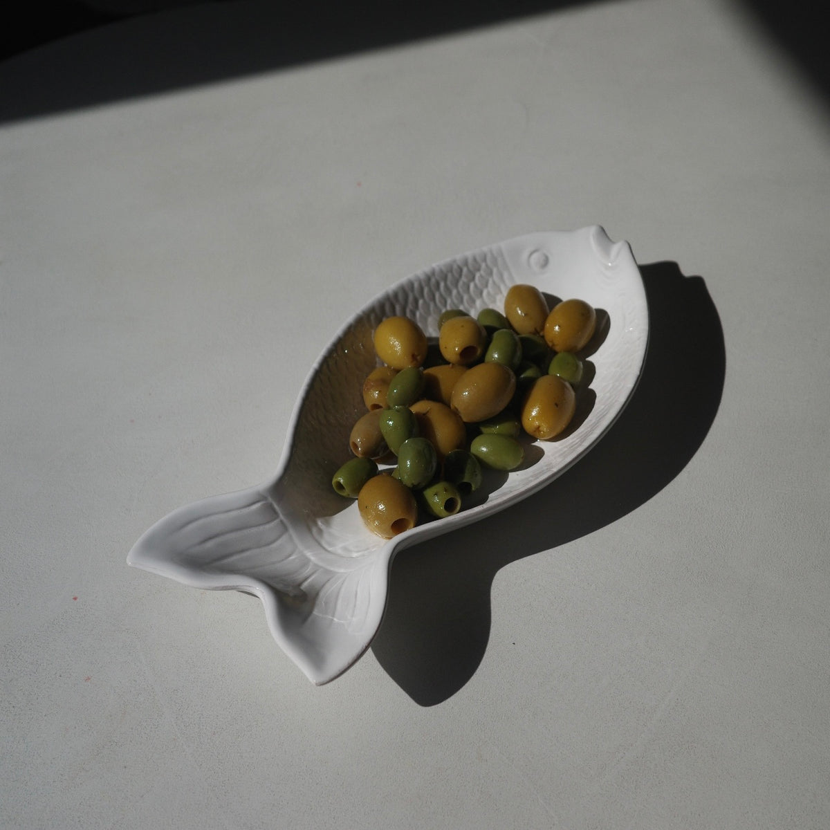 A white fish-shaped ceramic dish filled with green and yellow olives, photographed on a light gray surface with a strong cast shadow.