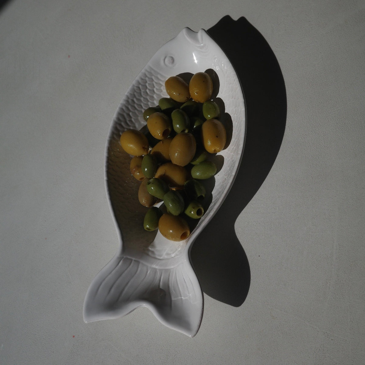 A white ceramic fish-shaped bowl filled with green and yellow olives, photographed on a light gray surface with a strong shadow.