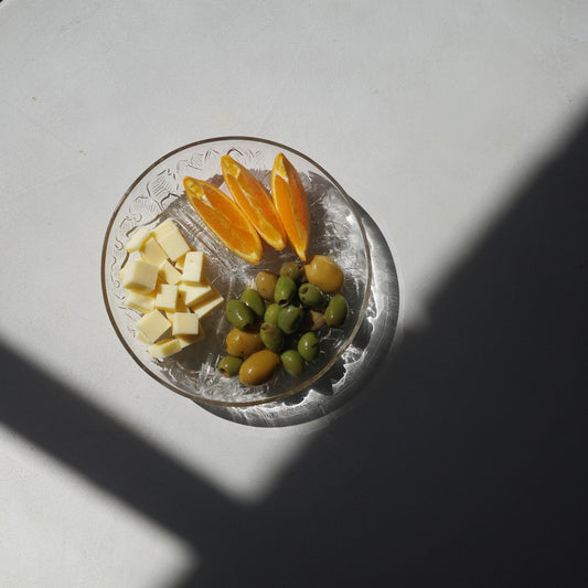 A clear glass plate holding cubed pale yellow cheese, orange slices, and green olives, photographed from above on a white surface with strong diagonal shadows.