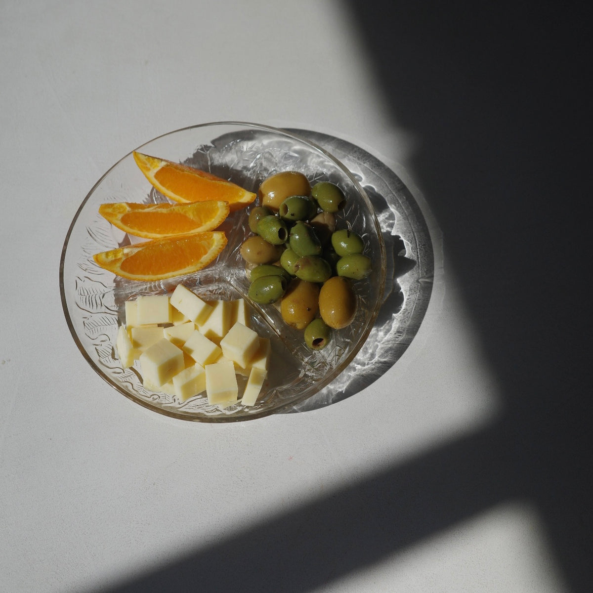 A clear glass plate with embossed floral detailing, holding cubed cheese, green and yellow olives, and orange slices, set on a white surface under direct sunlight with a strong shadow.