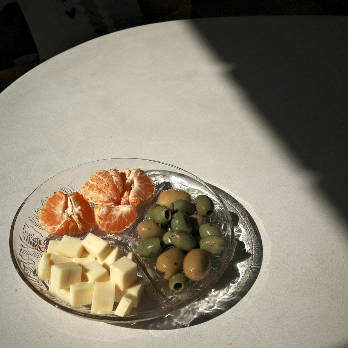A clear glass serving platter on a white table, filled with cubed pale yellow cheese, orange mandarin segments, and green and brown olives, under direct sunlight with a strong shadow.