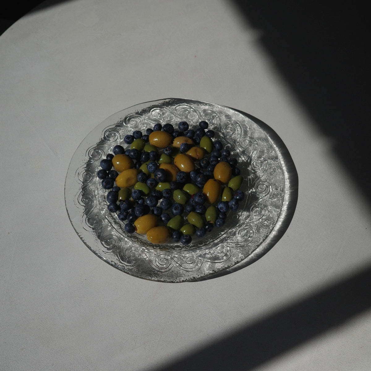 A clear glass plate with an embossed floral pattern, filled with blueberries and small yellow-green olives, set on a white surface under direct sunlight with strong shadows.