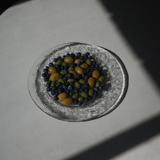 A clear glass plate with an embossed floral pattern, filled with blueberries and small yellow-green olives, set on a white surface under direct sunlight with strong shadows.