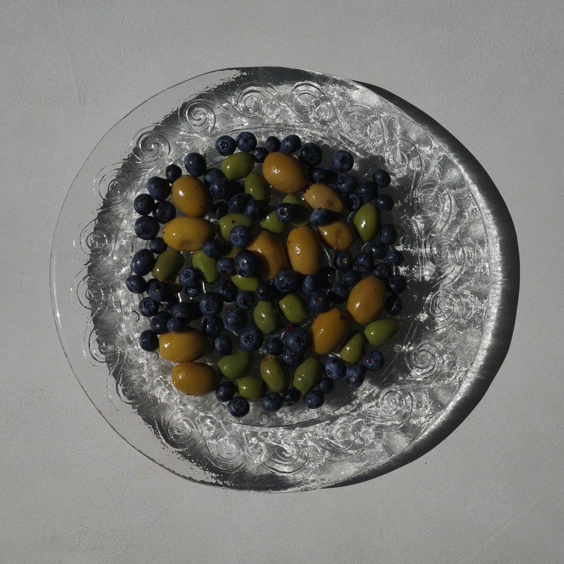 A top-down view of a clear glass plate with an embossed floral pattern, filled with blueberries, green olives, and yellow olives on a neutral light gray background.