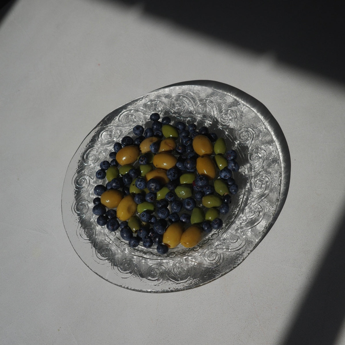 A top-down view of a clear glass plate with an embossed floral border, filled with blueberries and small yellow-green olives, set on a light gray surface with strong shadows.