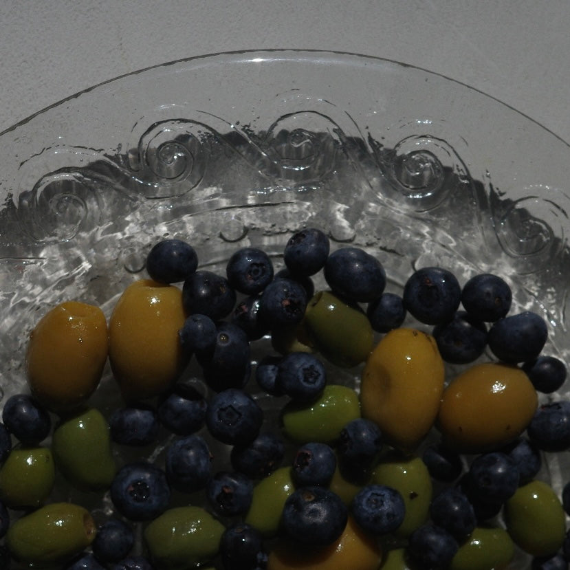 A close-up of mixed fruit in a clear glass bowl, including dark blue blueberries, yellow-orange yellow olives, and green green olives.