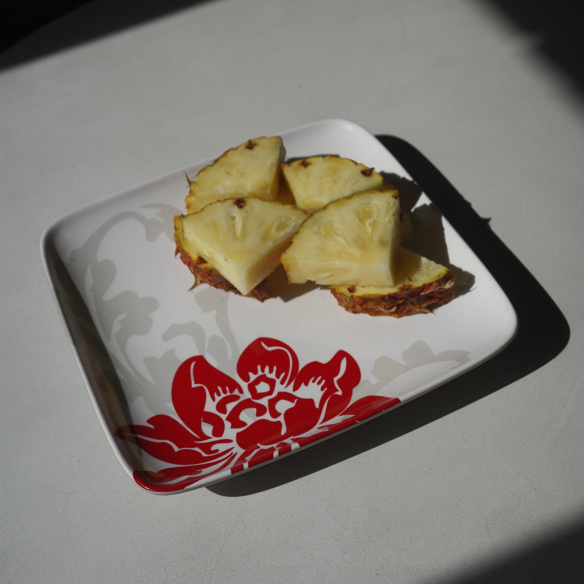 Four slices of pineapple resting on a square white  ceramic plate with a red floral pattern and gray scrollwork, placed on a light gray surface in direct sunlight.
