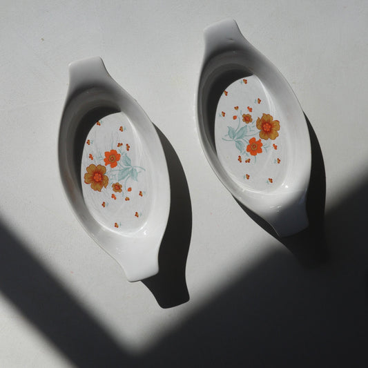 Two white ceramic oval baking dishes with orange and yellow floral patterns, photographed on a white surface with strong diagonal shadows.