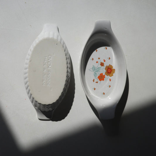Two white ceramic oven dishes shown on a white surface in strong sunlight, with one dish displaying its textured white bottom saying ovenproof and the other showing a floral pattern with orange flowers and green leaves.
