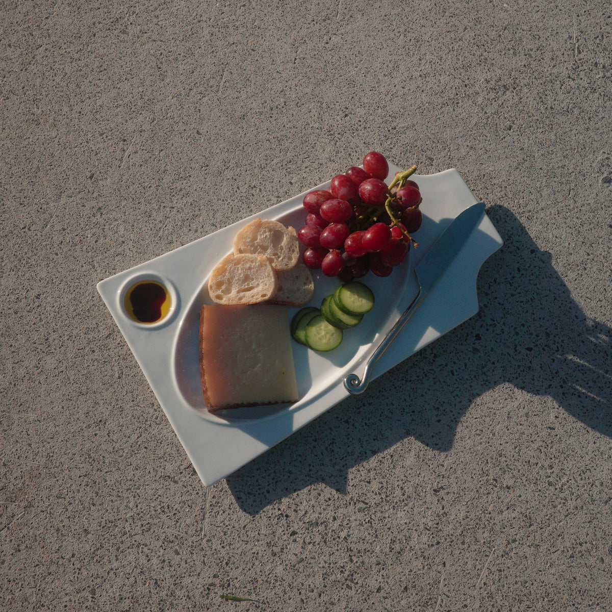 A white rectangular serving tray containing sliced bread, a piece of cheese, grapes, cucumber slices, and a small dipping sauce, placed on a gray concrete surface outdoors.