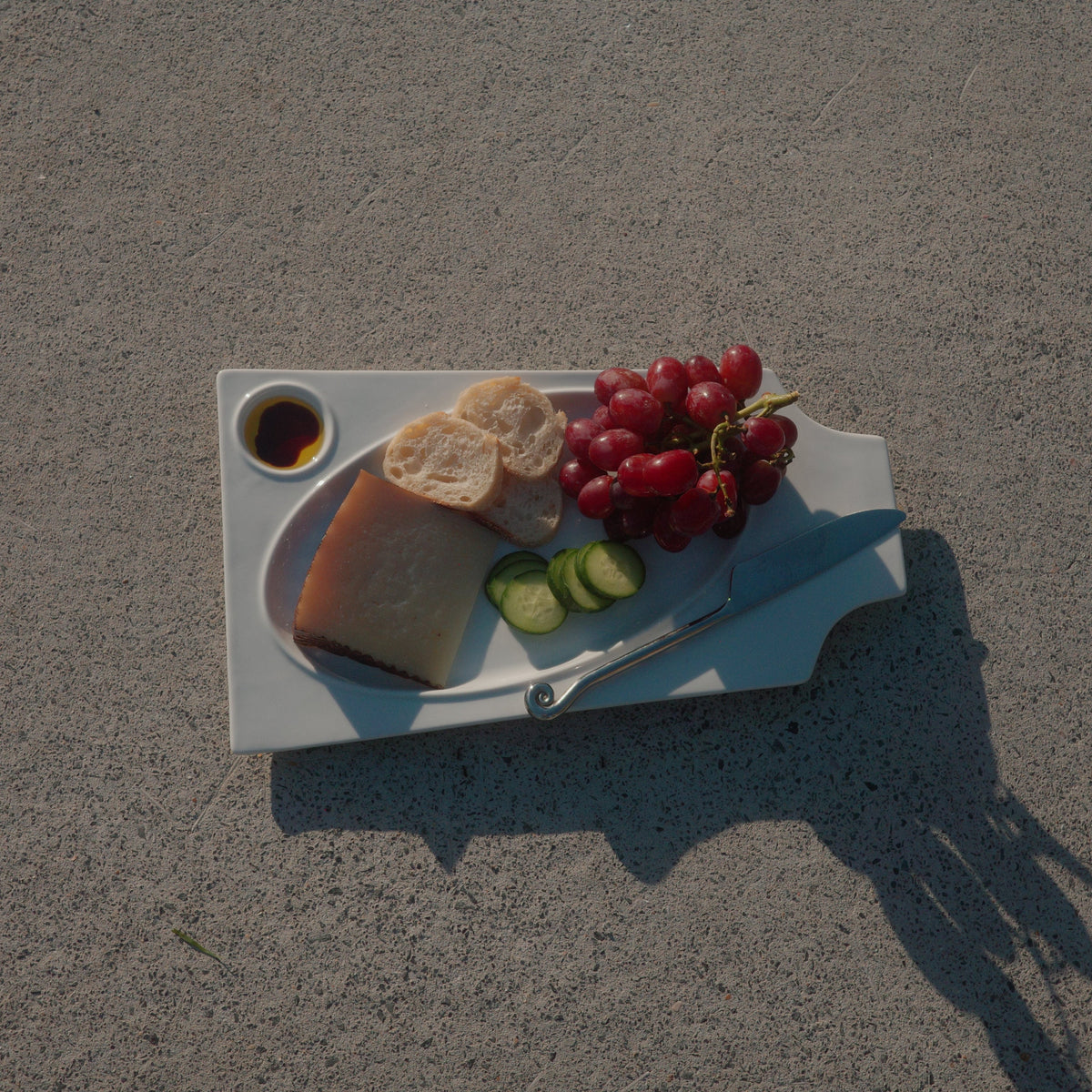 A white serving tray holding a wedge of cheese, sliced bread, red grapes, cucumber slices, and a small dipping sauce, placed on a gray paved surface with a strong shadow.