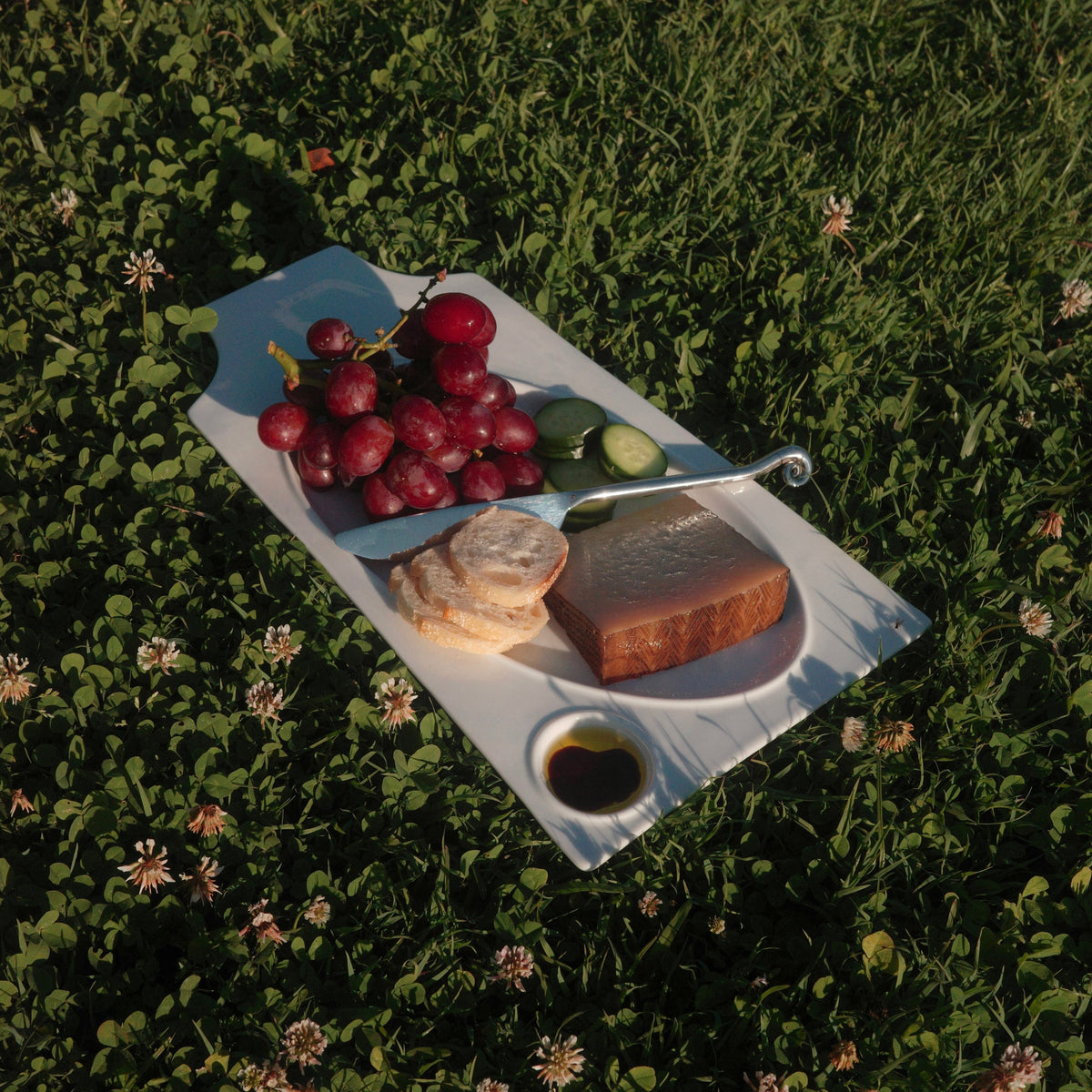 A white ceramic platter board laid on green grass, holding red grapes, a cucumber, bread, a slice of cheese, a small bowl of dressing.
