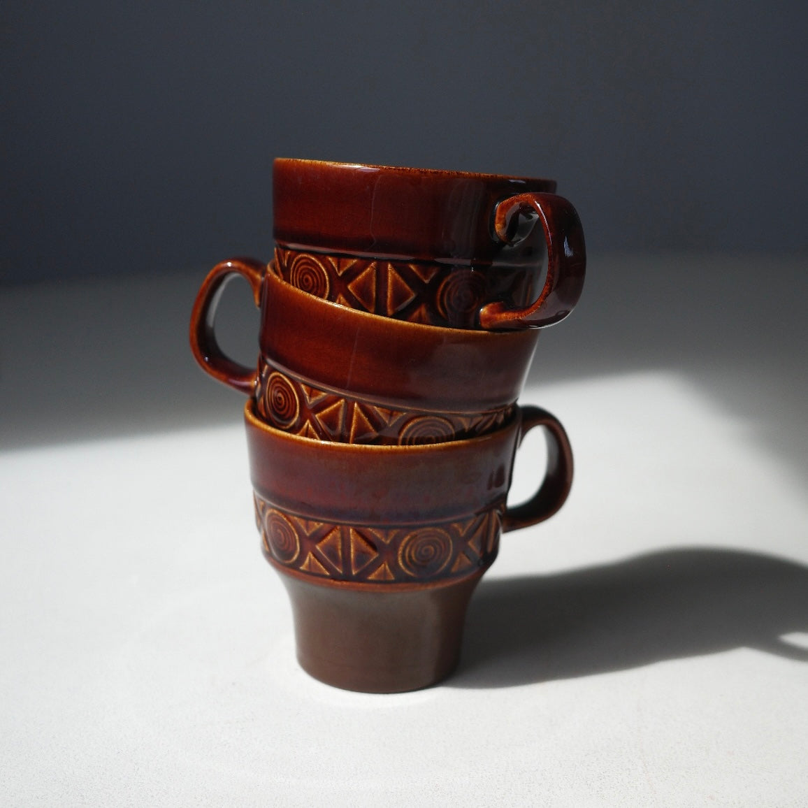 Three stacked ceramic coffee cups with dark brown handles and geometric patterns, photographed on a white surface with a dark gray background.