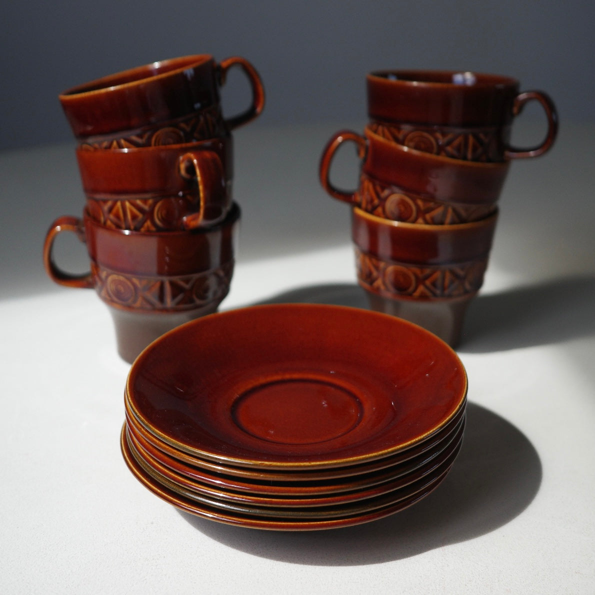 A set of vintage ceramic tableware in a glossy reddish-brown glaze, including four stacked cups with geometric embossed patterns and a stack of matching saucers on a light gray surface.