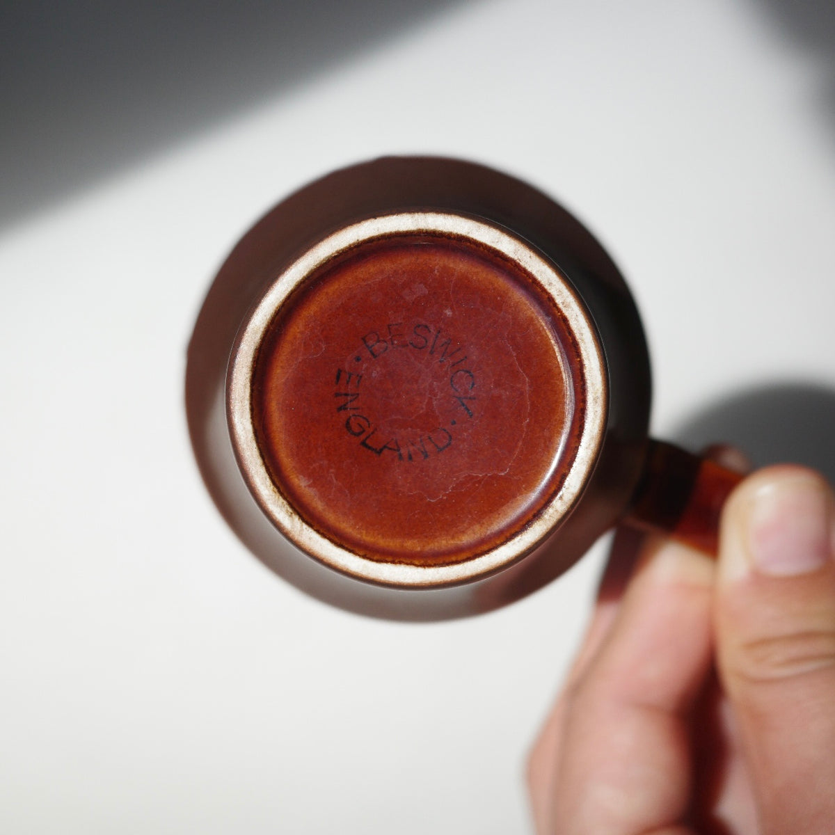 A close-up of a hand holding the handle of a brown ceramic mug, showing the bottom of the mug with a stamped brand name 