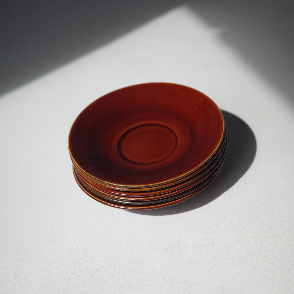 A stack of five ceramic plates in a glossy reddish-brown glaze, photographed on a light gray surface with a sharp diagonal shadow.