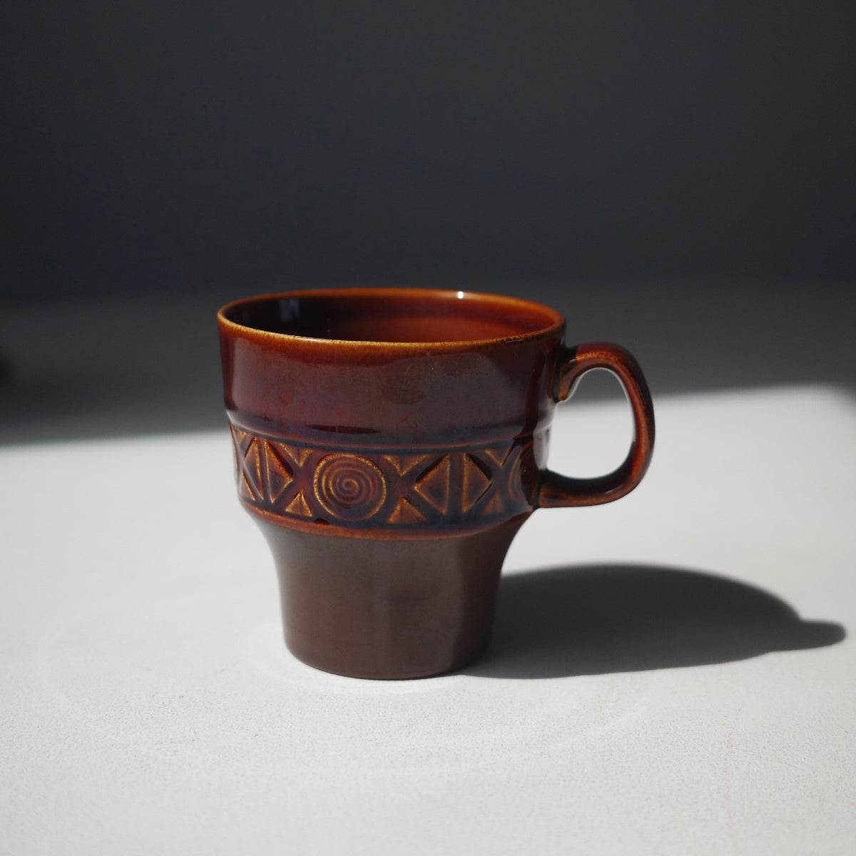 A brown ceramic coffee cup with a glossy finish and geometric patterns on the band, set against a dark gray background with a white surface.