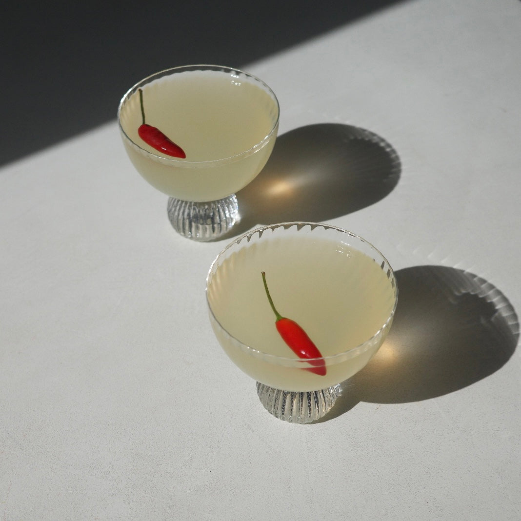 Two clear glass coupe-style cocktails glasses filled with a pale yellow liquid and each containing a small red chili pepper, photographed on a white surface with strong sunlight and shadows.