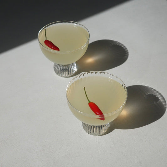 Two clear glass coupe-style cocktails glasses filled with a pale yellow liquid and each containing a small red chili pepper, photographed on a white surface with strong sunlight and shadows.