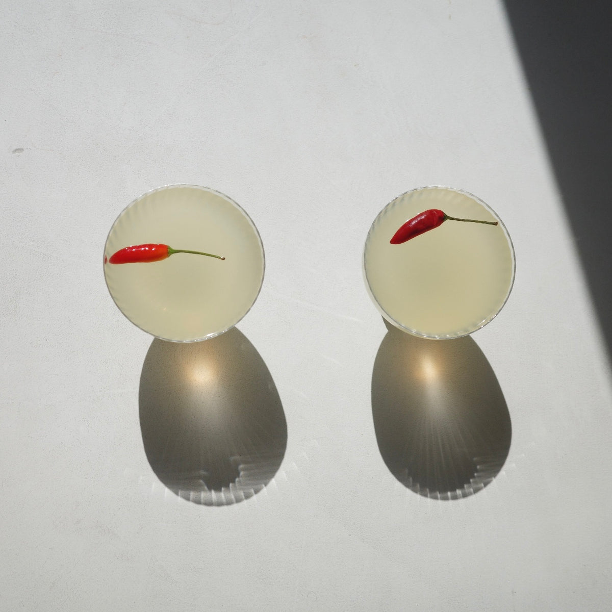 Two clear glass coupe-style cocktails glasses filled with a pale yellow liquid and each containing a small red chili pepper, photographed on a white surface with strong sunlight and shadows.