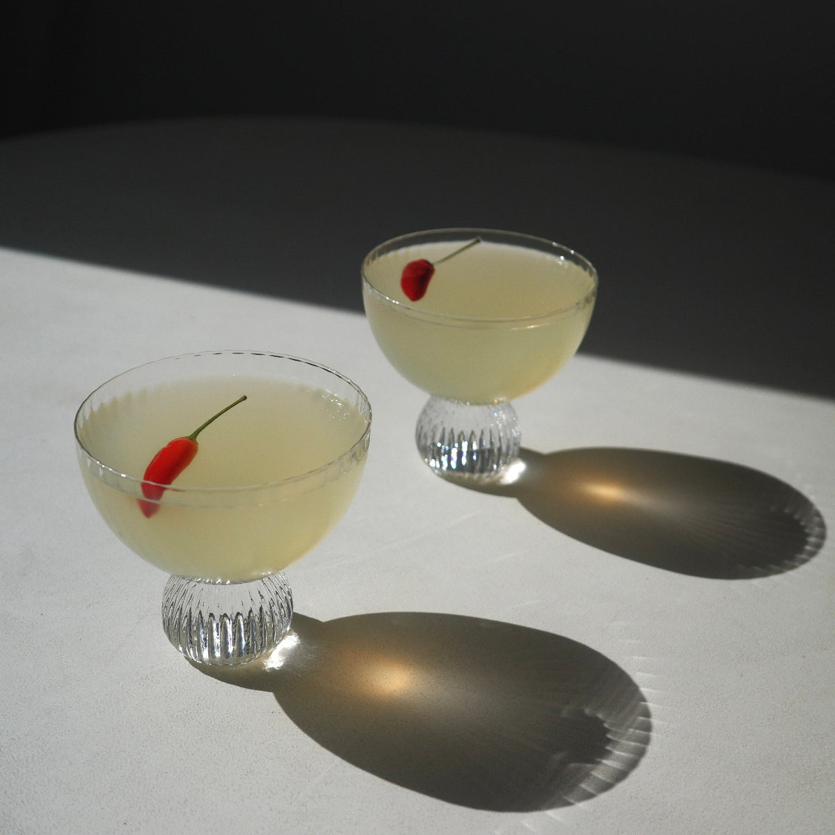 Two clear glass coupe-style cocktails glasses filled with a pale yellow liquid and each containing a small red chili pepper, photographed on a white surface with strong sunlight and shadows.