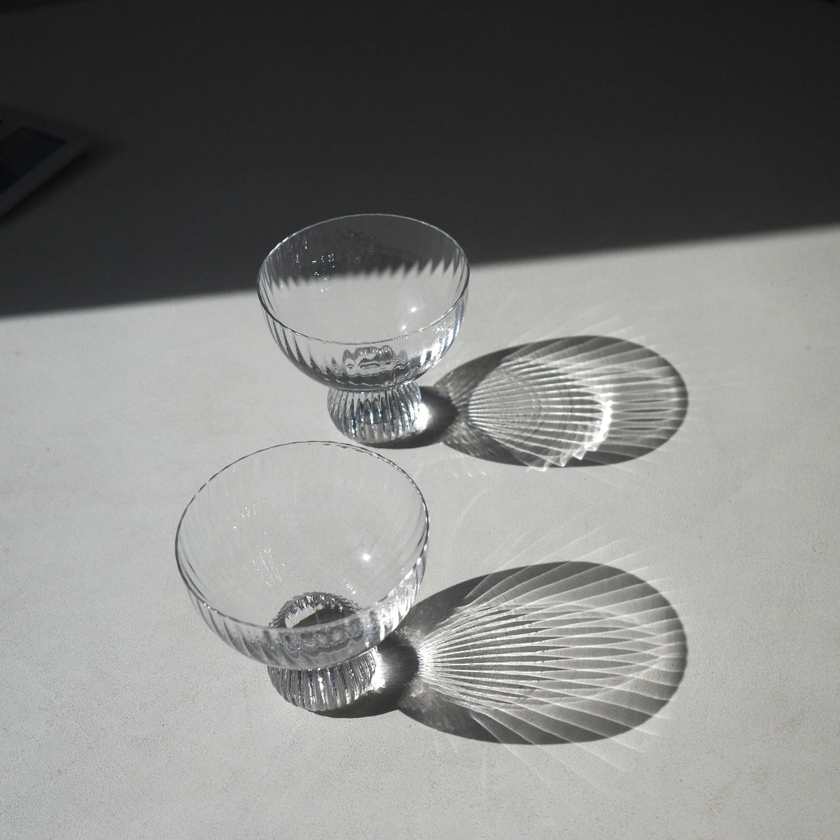 Two clear glass coupe-style cocktails glasses filled with a pale yellow liquid and each containing a small red chili pepper, photographed on a white surface with strong sunlight and shadows.
