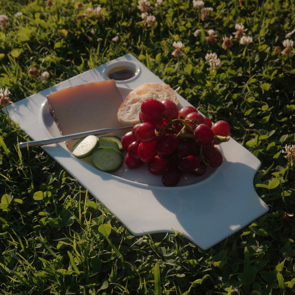 A white ceramic serving platter with cutouts, holding a slice of cheese, bread, grapes, and cucumber slices, placed on green grass outdoors.