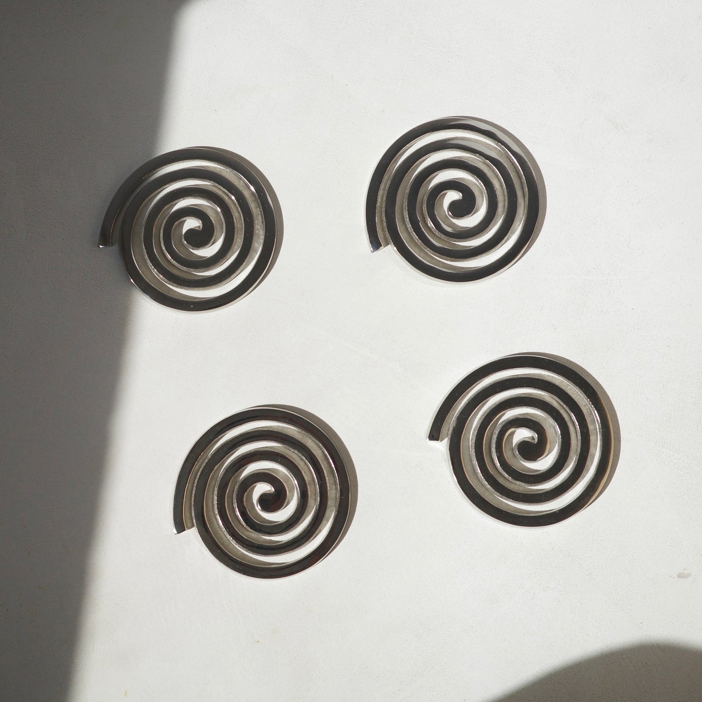 Four spiral-shaped metallic coasters arranged in a square on a white surface with soft shadows.