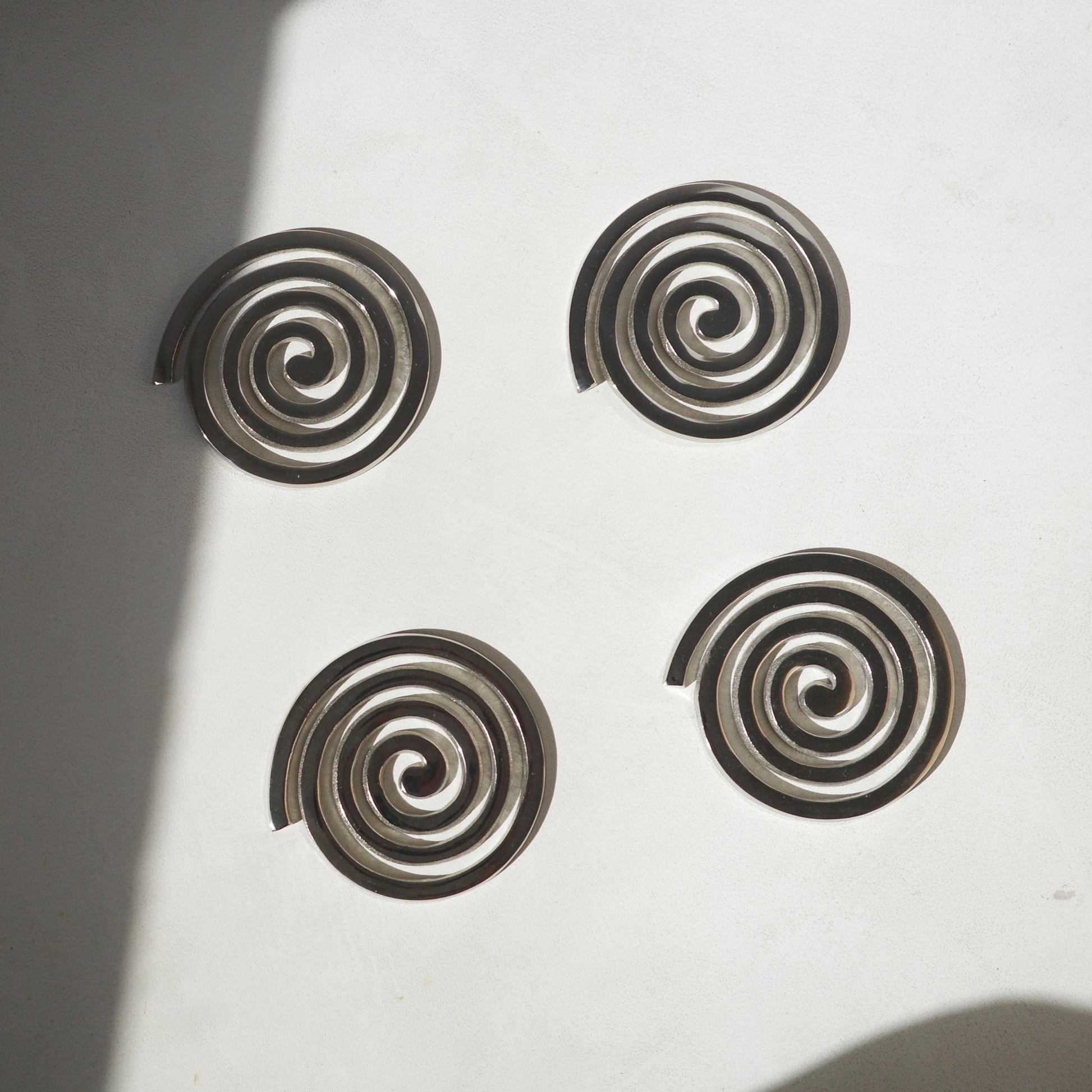 Four spiral-shaped metallic coasters arranged in a square on a white surface with soft shadows.